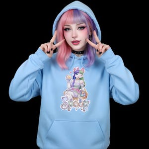 Pode incluir: Um moletom azul claro com um bolso frontal e um capuz com cordão. O moletom apresenta um desenho animado de um coelho com a palavra "Spoop" em uma fonte decorativa. A pessoa na imagem está usando um choker preto e tem cabelo rosa e roxo.