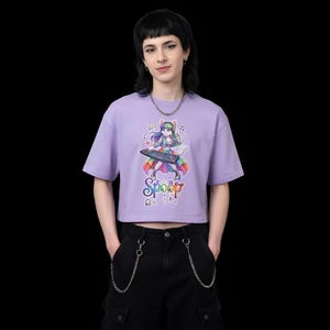 Pode incluir: Um top cropped lilás com um gráfico colorido de um personagem de desenho animado tocando um teclado. O personagem tem uma cauda de arco-íris e é cercado por notas musicais. A palavra "Spoop" está escrita em uma fonte gotejante.