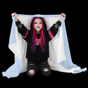 Pode incluir: Uma manta com capuz com borda azul claro e interior branco é levantada. A manta está sendo levantada por uma pessoa vestindo um moletom preto com detalhes em rosa e jeans pretos rasgados. A pessoa tem cabelo rosa e roxo.