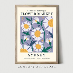 May include: A framed art print featuring a floral design with white and yellow daisies on a lavender background. The print includes the text "FLOWER MARKET SYDNEY" and "Collection Botanique". The frame is a light wood color.