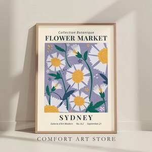 May include: A framed art print featuring a floral design with white and yellow daisies on a lavender background. The print includes the text "Flower Market Sydney" and "Comfort Art Store". The artwork is in a light wooden frame.
