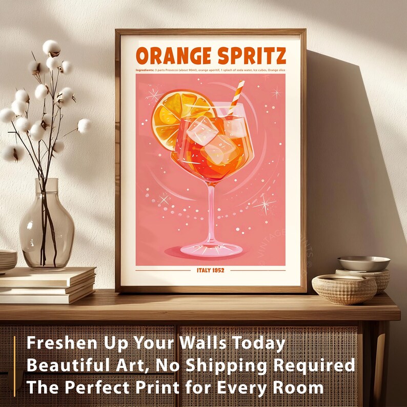 Puede incluir: Un cuadro enmarcado con una ilustraci&oacute;n del c&oacute;ctel "Orange Spritz". La obra de arte muestra un c&oacute;ctel naranja brillante con cubitos de hielo y una rodaja de naranja, sobre un fondo rosa. El texto "ITALY 1952" est&aacute; en la parte inferior. El cuadro se exhibe sobre una superficie de madera.