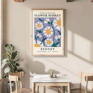 May include: A framed poster with a floral design, featuring white daisies with yellow centers on a lavender background. The poster reads "FLOWER MARKET SYDNEY" in a vintage style. The poster is displayed in a room with a table and chairs.