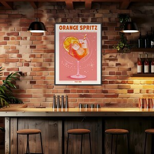 Puede incluir: Un p&oacute;ster enmarcado con las palabras "ORANGE SPRITZ" en la parte superior. El p&oacute;ster presenta un gr&aacute;fico de un c&oacute;ctel de naranja con cubitos de hielo y una pajita. El fondo es de color rosa claro. El p&oacute;ster est&aacute; colgado en una pared de ladrillo sobre una barra.