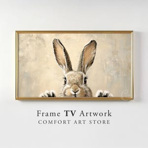 May include: A framed print of a brown rabbit peeking out from behind a beige background. The rabbit has long ears and a fluffy tail. The frame is gold and has a simple design.