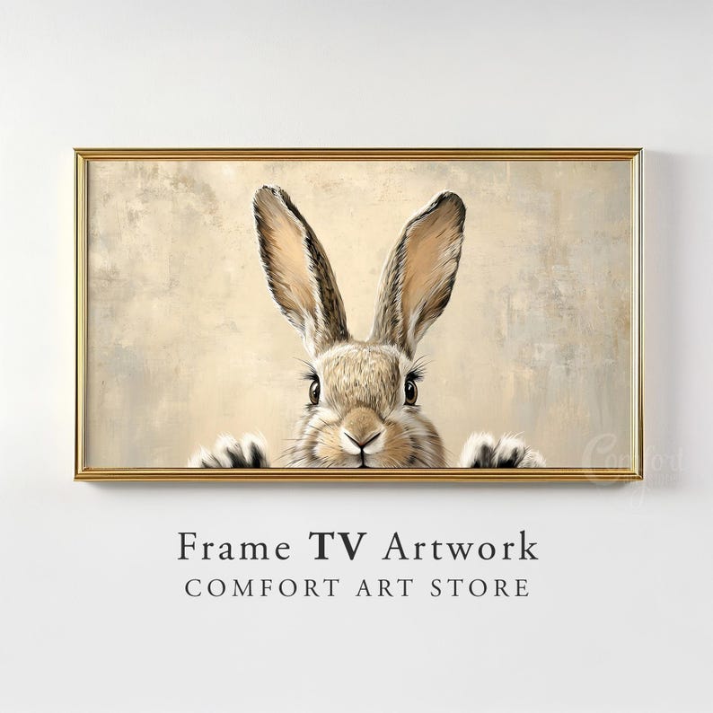 May include: A framed print of a brown rabbit peeking out from behind a beige background. The rabbit has long ears and a fluffy tail. The frame is gold and has a simple design.