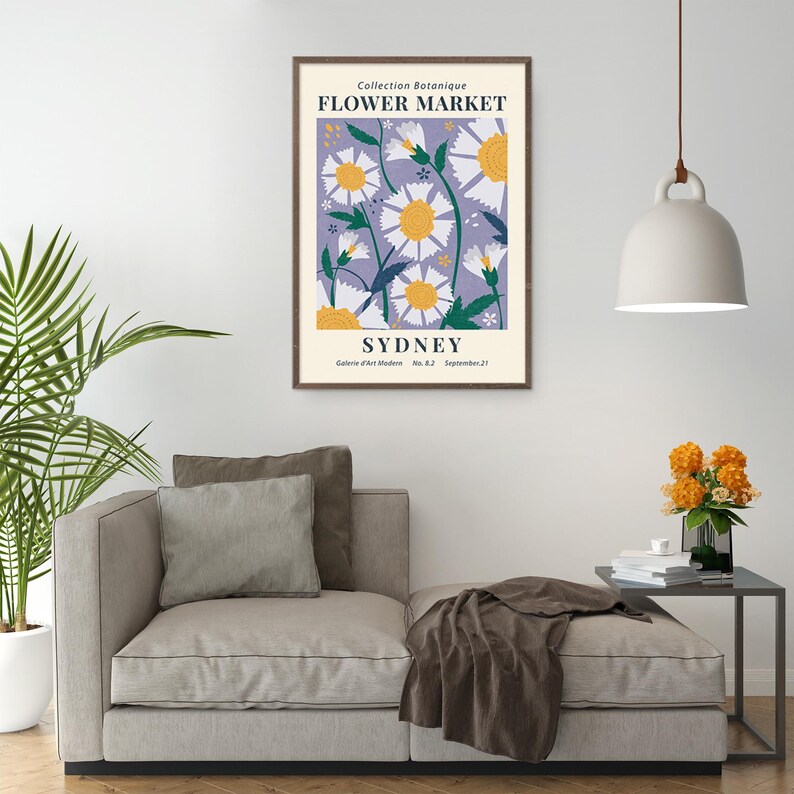 May include: A framed art print featuring a floral design with white daisies and yellow centers against a lavender background. The print is labeled "Flower Market Sydney" and is displayed above a beige sofa with pillows.