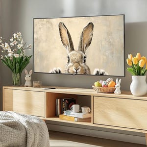 May include: A light brown rabbit with long ears peeks out from behind a television screen. The rabbit is looking directly at the viewer. The television is on a wooden entertainment center with a vase of yellow flowers and a bowl of colorful Easter eggs.