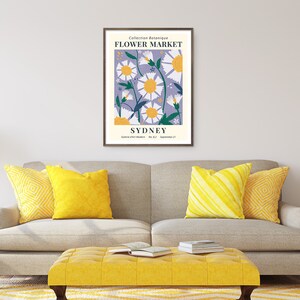 May include: A framed art print featuring a floral design with daisies and the text "FLOWER MARKET SYDNEY." The artwork has a lavender background with white and yellow flowers. The print is displayed above a gray sofa with yellow pillows and a yellow ottoman.