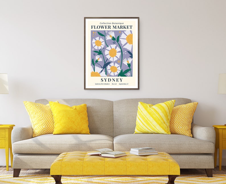 May include: A framed art print featuring a floral design with daisies and the text "FLOWER MARKET SYDNEY." The artwork has a lavender background with white and yellow flowers. The print is displayed above a gray sofa with yellow pillows and a yellow ottoman.