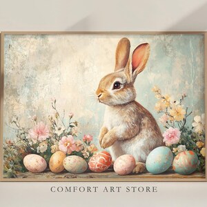 Puede incluir: Una pintura de un conejo marr&oacute;n sentado entre flores rosas y blancas con acentos azul pastel y amarillo. El conejo est&aacute; rodeado de huevos de Pascua coloridos.