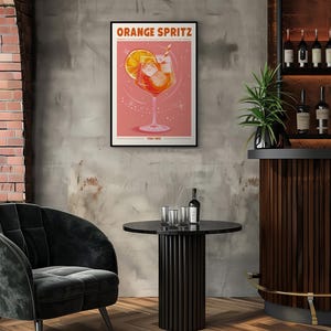 Puede incluir: Un p&oacute;ster enmarcado con las palabras "ORANGE SPRITZ" presenta una ilustraci&oacute;n de c&oacute;ctel con una rodaja de naranja y cubitos de hielo. El p&oacute;ster se exhibe en un ambiente de bar con una mesa negra, una silla de terciopelo y una estanter&iacute;a de licores.