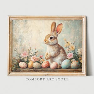 Puede incluir: Una pintura enmarcada de un conejo marr&oacute;n sentado entre un lecho de flores rosas y blancas. El conejo est&aacute; rodeado de huevos de Pascua coloridos.