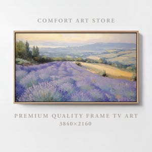 May include: A framed landscape painting of a field of purple lavender flowers in a valley. The painting is set against a backdrop of rolling hills and a hazy sky. The painting is framed in a light wood frame.