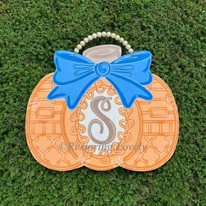 May include: A pumpkin-shaped wooden door hanger featuring an orange surface with a white and orange lattice pattern, a large blue bow, a central white oval with a brown letter S, and a beaded hanger. The text Restoring Lovely is visible.
