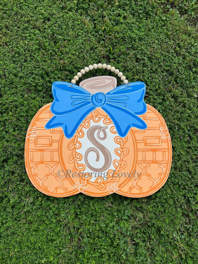 May include: A pumpkin-shaped wooden door hanger featuring an orange surface with a white and orange lattice pattern, a large blue bow, a central white oval with a brown letter S, and a beaded hanger. The text Restoring Lovely is visible.