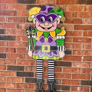 May include: A purple and green jester with a yellow hat and a gold belt. The jester is holding a scepter with a fleur-de-lis on top. The jester is wearing black and white striped stockings and black shoes. The jester is standing in front of a brick wall.