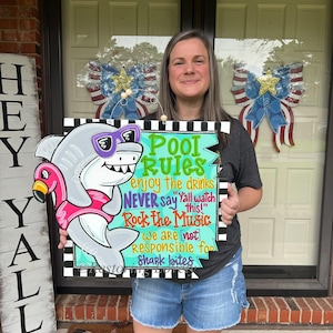 May include: A wooden sign with a cartoon shark wearing sunglasses and a flamingo floatie. The sign reads "Pool Rules" with phrases like "enjoy the drinks" and "Rock the Music." The sign is held by a person in front of a door.