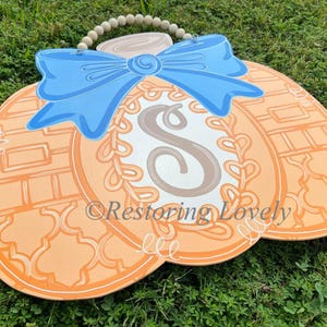 May include: A flat, pumpkin-shaped wooden door hanger in shades of orange, featuring a large light blue bow at the top, a central oval with the letter S, and a beaded hanging cord. The text Restoring Lovely is printed across the center.