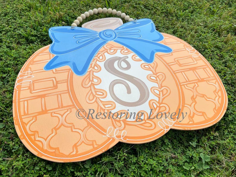 May include: A flat, pumpkin-shaped wooden door hanger in shades of orange, featuring a large light blue bow at the top, a central oval with the letter S, and a beaded hanging cord. The text Restoring Lovely is printed across the center.