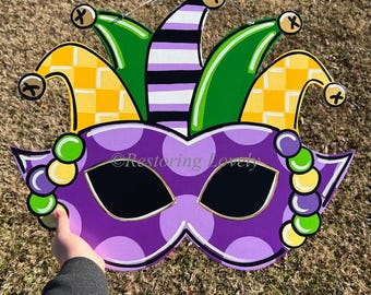 May include: Purple and green Mardi Gras mask with a jester hat. The mask has polka dots and beads. The hat has green and yellow checkered patterns and stripes.