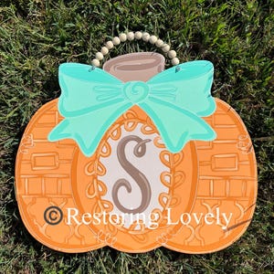 May include: A pumpkin-shaped wooden door hanger featuring an orange textured surface, a large teal bow, and a central oval containing the letter S. A string of light-colored wooden beads serves as a hanger. The text Restoring Lovely is visible.
