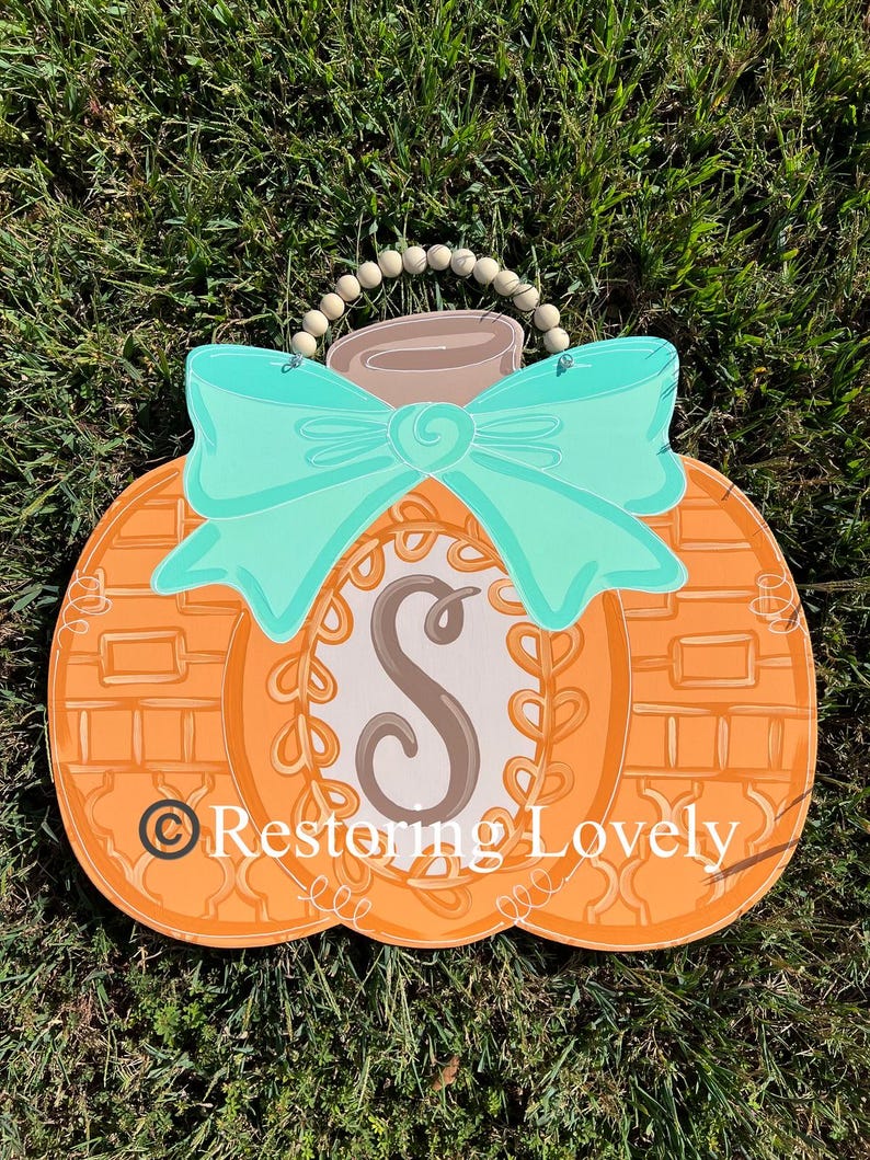 May include: A pumpkin-shaped wooden door hanger featuring an orange textured surface, a large teal bow, and a central oval containing the letter S. A string of light-colored wooden beads serves as a hanger. The text Restoring Lovely is visible.