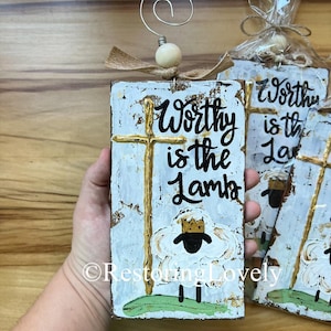 May include: A rustic, rectangular wooden sign with the text "Worthy is the Lamb" and a gold cross and sheep illustration. The sign is painted white with distressed accents and has a wire clip and wooden bead attached.