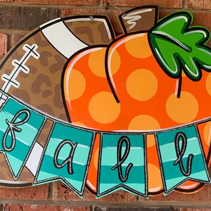 May include: A wooden door hanger decorated with a football and a pumpkin. The pumpkin is orange with white polka dots and a green stem. The football is brown with a leopard print pattern and white laces. A teal blue banner with the word "fall" in black cursive letters hangs below the football and pumpkin.