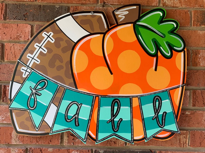 May include: A wooden door hanger decorated with a football and a pumpkin. The pumpkin is orange with white polka dots and a green stem. The football is brown with a leopard print pattern and white laces. A teal blue banner with the word "fall" in black cursive letters hangs below the football and pumpkin.