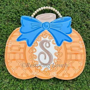 May include: A pumpkin-shaped wooden door hanger featuring a bright blue bow at the top, a central white oval with a brown letter S, and a decorative vine border. The pumpkin has orange sections with white line patterns and a wooden bead hanger.