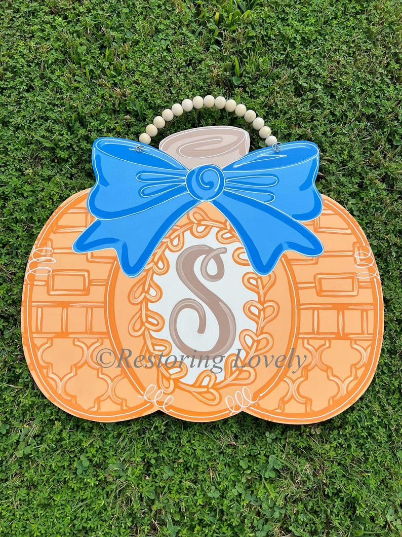 May include: A pumpkin-shaped wooden door hanger featuring a bright blue bow at the top, a central white oval with a brown letter S, and a decorative vine border. The pumpkin has orange sections with white line patterns and a wooden bead hanger.
