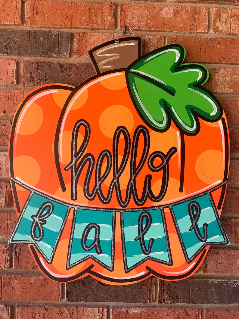 May include: Orange pumpkin-shaped wooden door hanger with white polka dots and a green leaf. The pumpkin has the words "hello fall" written on it in black with a silver glitter outline.