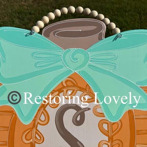 May include: A wooden door hanger shaped like a pumpkin, painted orange with a textured pattern, featuring a large teal bow, a brown stem, and a wooden bead hanger. The center displays a circular design with the letter S and the text Restoring Lovely.