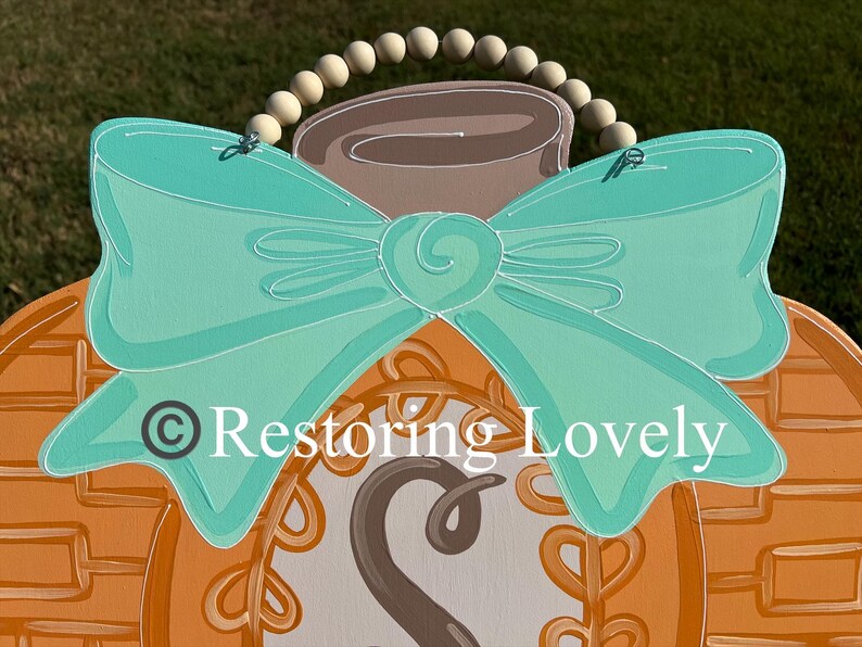 May include: A wooden door hanger shaped like a pumpkin, painted orange with a textured pattern, featuring a large teal bow, a brown stem, and a wooden bead hanger. The center displays a circular design with the letter S and the text Restoring Lovely.