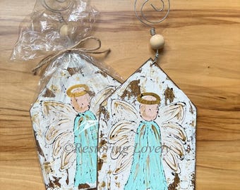 May include: Two angel-themed ornaments with a distressed white and brown finish. Each ornament features a hand-painted angel in a light blue robe with a gold halo. A wire spiral and wooden bead are attached to the top.