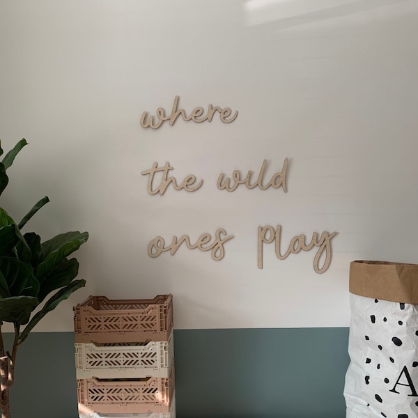 May include: Wooden wall decor with the words "where the wild ones play" in a cursive font. The decor is mounted on a white wall with a light blue wall below. There are four stacked plastic storage crates in front of the wall. The crates are beige and white. There are two books on top of the crates. A brown paper bag with black polka dots is on the right side of the image.