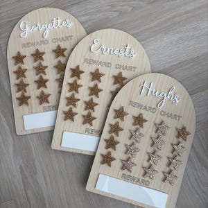 May include: Three wooden reward charts with gold glitter stars. The charts are labelled "George's Reward Chart", "Ernest's Reward Chart", and "Hugh's Reward Chart".