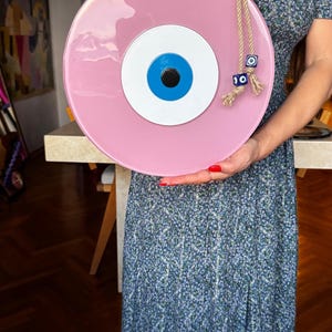 K&ouml;nnte beinhalten: Ein gro&szlig;es, rundes, rosa dekoratives Auge mit blauer Iris, wei&szlig;er Mitte und schwarzer Pupille. Das Auge ist an einem Seil aufgeh&auml;ngt und mit blauen und silbernen Perlen verziert. Ein dekorativer Artikel f&uuml;r die Heimdekoration.