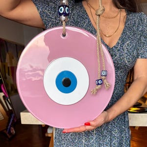 K&ouml;nnte beinhalten: Ein gro&szlig;er, runder, rosa Evil Eye-Wandbehang. Das Auge hat eine blaue Iris, einen wei&szlig;en Rand und eine schwarze Pupille. Es wird von einem Seil gehalten und mit blauen und wei&szlig;en Perlen verziert. Ein dekoratives St&uuml;ck f&uuml;r Schutz und Gl&uuml;ck.