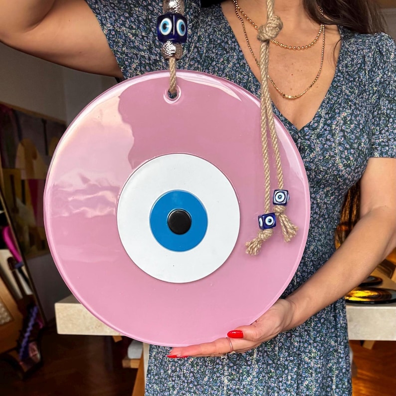 K&ouml;nnte beinhalten: Ein gro&szlig;er, runder, rosa Evil Eye-Wandbehang. Das Auge hat eine blaue Iris, einen wei&szlig;en Rand und eine schwarze Pupille. Es wird von einem Seil gehalten und mit blauen und wei&szlig;en Perlen verziert. Ein dekoratives St&uuml;ck f&uuml;r Schutz und Gl&uuml;ck.