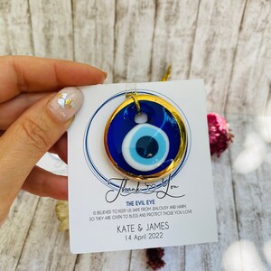 K&ouml;nnte beinhalten: Ein blauer und goldener b&ouml;ser Blick-Anh&auml;nger mit einem wei&szlig;en Zentrum. Der Anh&auml;nger befindet sich auf einer wei&szlig;en Karte mit dem Text "Thank You" in Kursivschrift, "The Evil Eye" in Gro&szlig;buchstaben und "IS BELIEVED TO KEEP US SAFE FROM JEALOUSY AND HARM SO THEY ARE GIVEN TO BLESS AND PROTECT THOSE YOU LOVE KATE & JAMES 14 April 2022".