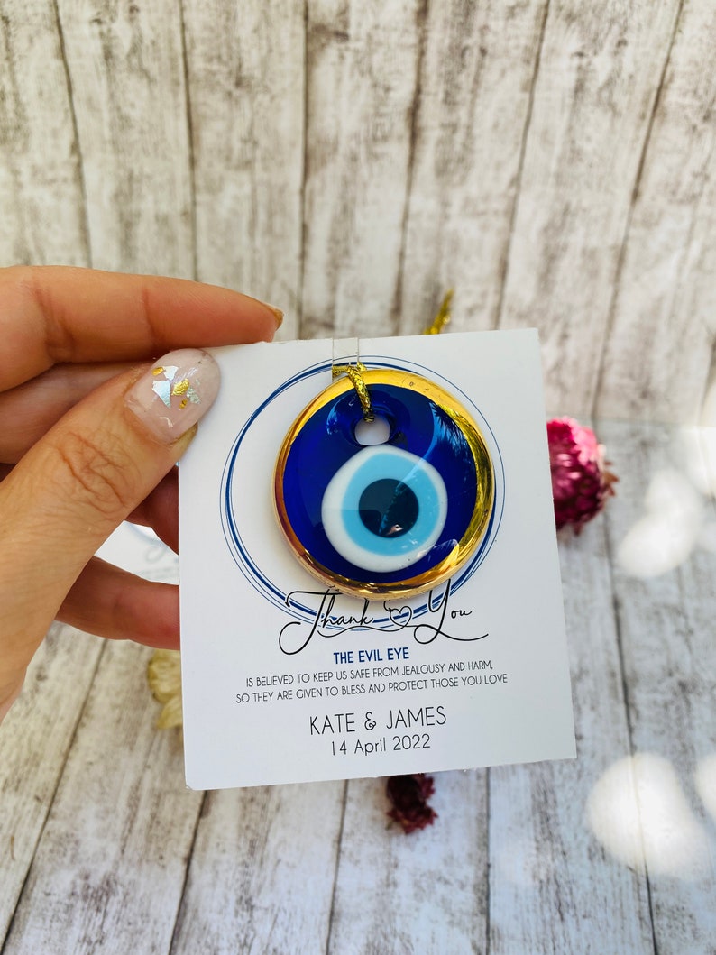 K&ouml;nnte beinhalten: Ein blauer und goldener b&ouml;ser Blick-Anh&auml;nger mit einem wei&szlig;en Zentrum. Der Anh&auml;nger befindet sich auf einer wei&szlig;en Karte mit dem Text "Thank You" in Kursivschrift, "The Evil Eye" in Gro&szlig;buchstaben und "IS BELIEVED TO KEEP US SAFE FROM JEALOUSY AND HARM SO THEY ARE GIVEN TO BLESS AND PROTECT THOSE YOU LOVE KATE & JAMES 14 April 2022".
