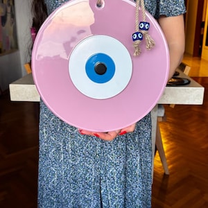 K&ouml;nnte beinhalten: Ein gro&szlig;er, runder, rosa Wandbehang mit einem zentralen Augendesign. Das Auge hat eine wei&szlig;e Iris, eine blaue Pupille und eine schwarze Mitte. Es wird von einem Naturseil mit dekorativen blauen Augenperlen gehalten.