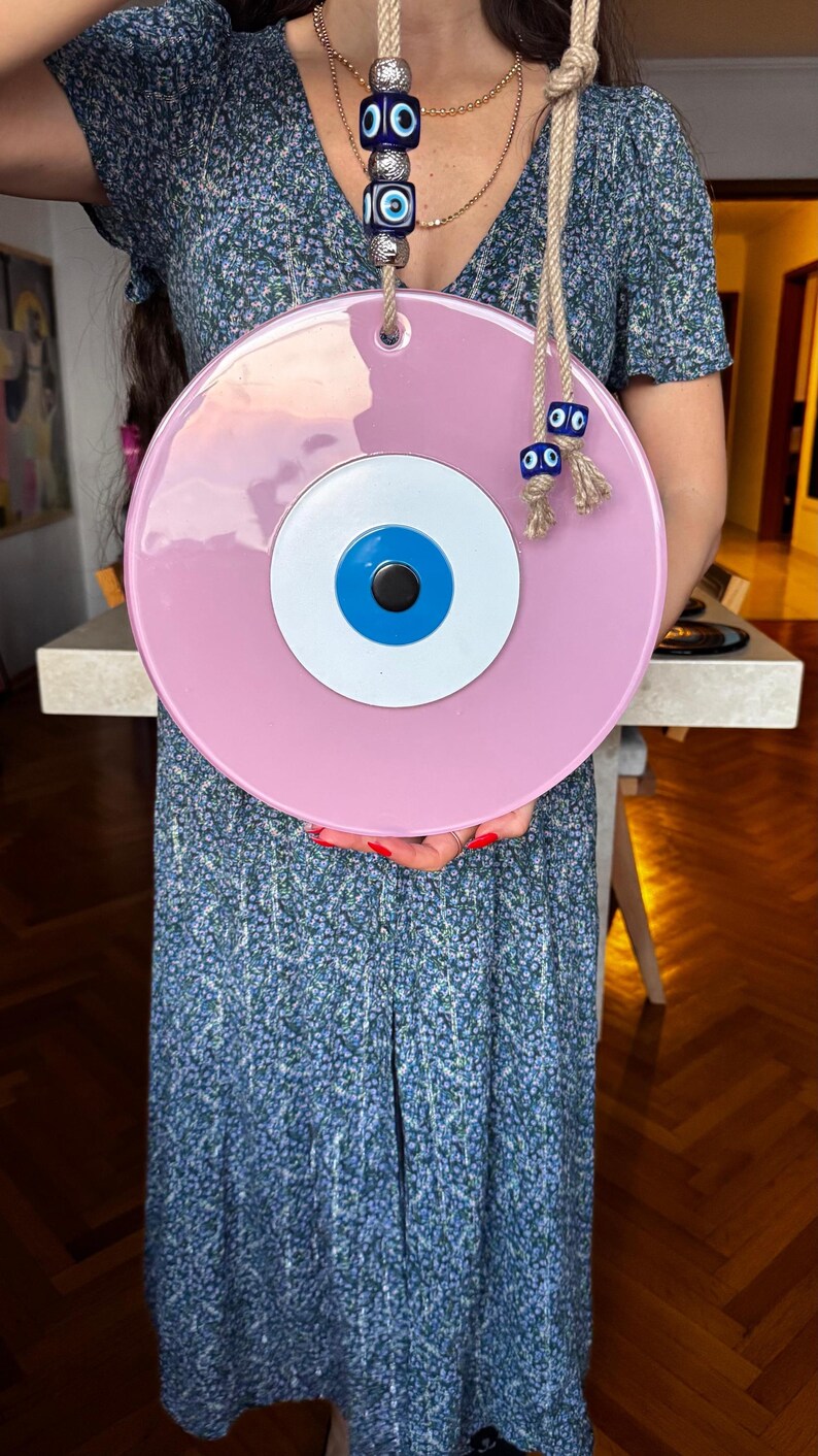 K&ouml;nnte beinhalten: Ein gro&szlig;er, runder, rosa Wandbehang mit einem zentralen Augendesign. Das Auge hat eine wei&szlig;e Iris, eine blaue Pupille und eine schwarze Mitte. Es wird von einem Naturseil mit dekorativen blauen Augenperlen gehalten.