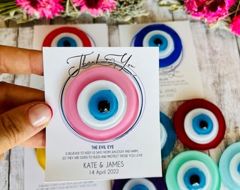 May include: Thank You card with a pink, white, and blue evil eye charm. The card says "The Evil Eye is believed to keep us safe from jealousy and harm so they are given to bless and protect those you love. Kate & James 14 April 2022."