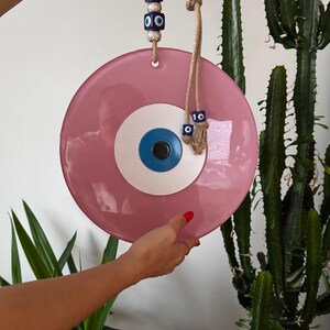 K&ouml;nnte beinhalten: Ein rosa Wandbehang mit b&ouml;sem Blick und blau-wei&szlig;em Augendesign. Das dekorative St&uuml;ck wird von einem Naturseil gehalten und hat blaue und silberne Perlen. Ein einzigartiger Einrichtungsgegenstand.