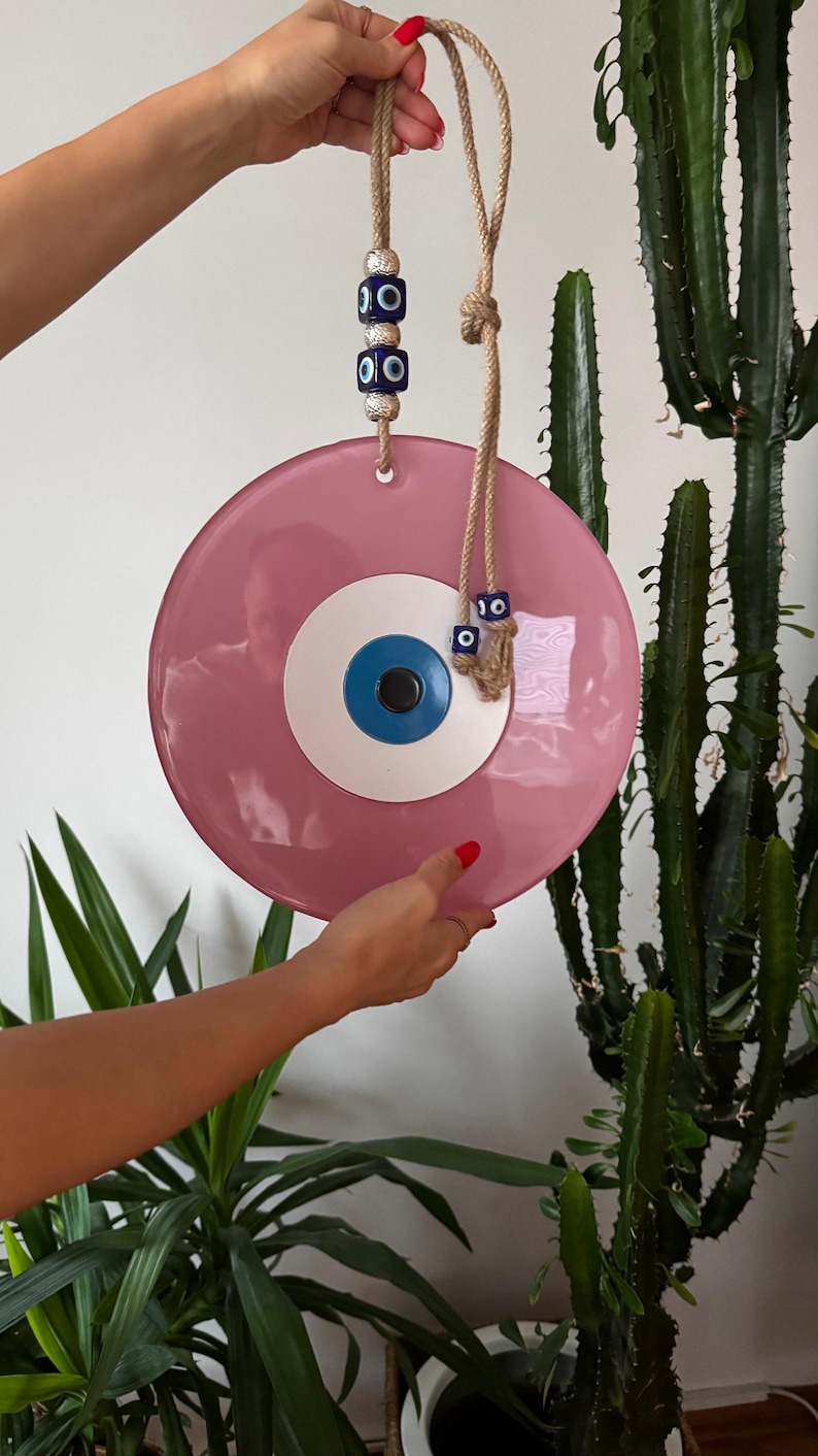 K&ouml;nnte beinhalten: Ein rosa Wandbehang mit b&ouml;sem Blick und blau-wei&szlig;em Augendesign. Das dekorative St&uuml;ck wird von einem Naturseil gehalten und hat blaue und silberne Perlen. Ein einzigartiger Einrichtungsgegenstand.