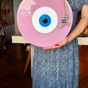 K&ouml;nnte beinhalten: Ein gro&szlig;es, rundes, rosa dekoratives Auge mit blauer Iris, wei&szlig;er Mitte und schwarzer Pupille. Das Auge ist an einem Seil aufgeh&auml;ngt und mit blauen und silbernen Perlen verziert. Ein dekorativer Artikel f&uuml;r die Heimdekoration.