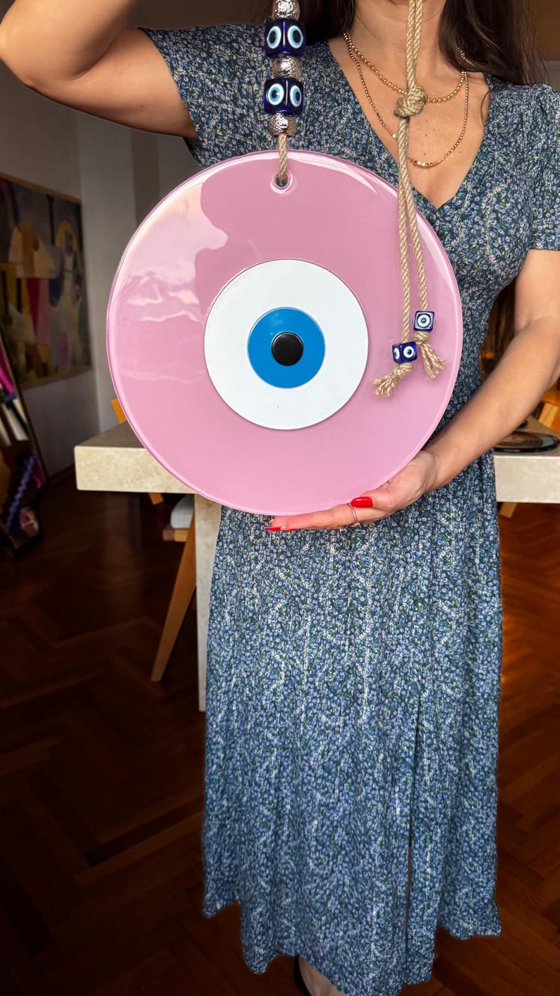 K&ouml;nnte beinhalten: Ein gro&szlig;es, rundes, rosa dekoratives Auge mit blauer Iris, wei&szlig;er Mitte und schwarzer Pupille. Das Auge ist an einem Seil aufgeh&auml;ngt und mit blauen und silbernen Perlen verziert. Ein dekorativer Artikel f&uuml;r die Heimdekoration.